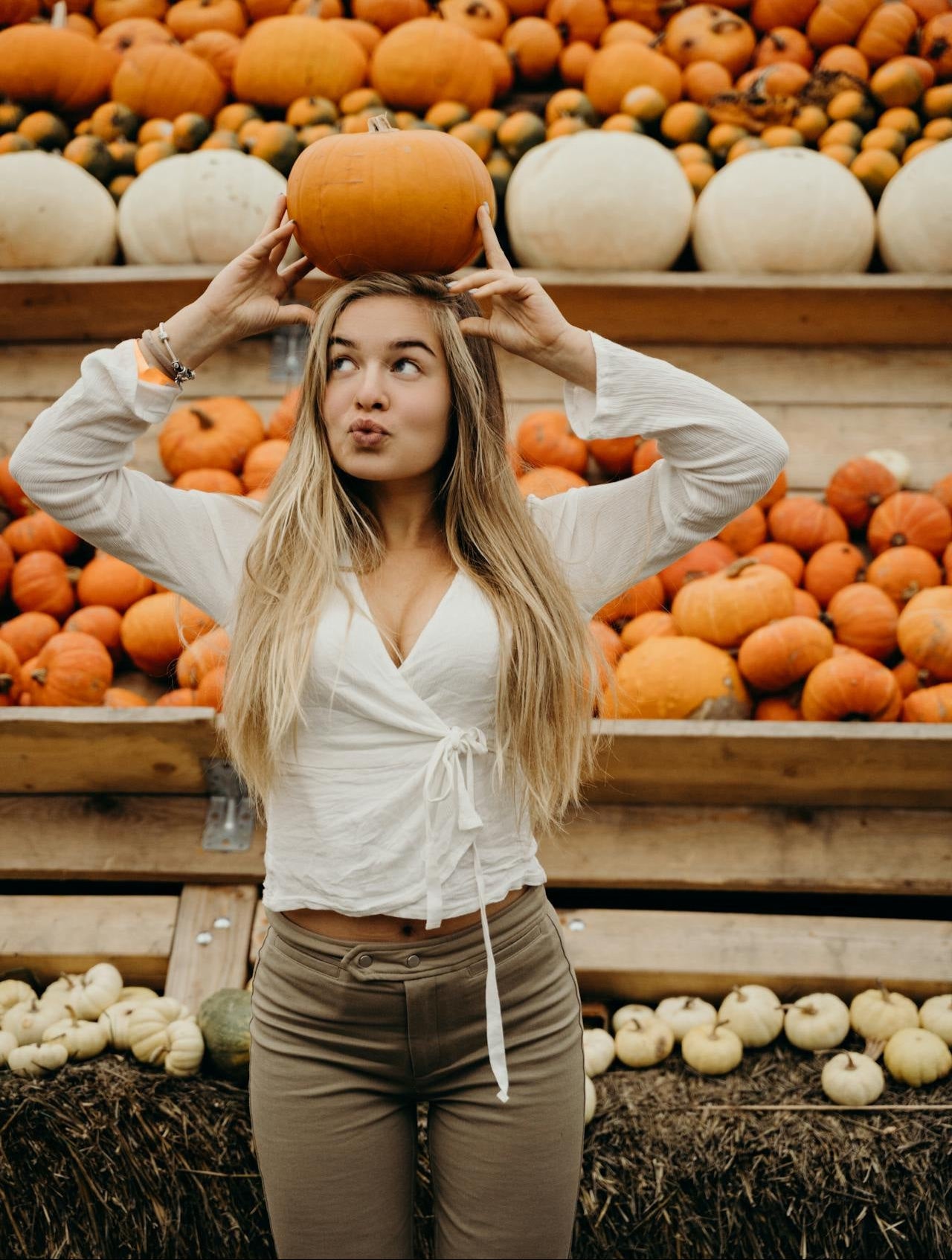 Woman holding a pumpkin above her head in front of stacked pumpkins and gourds.
