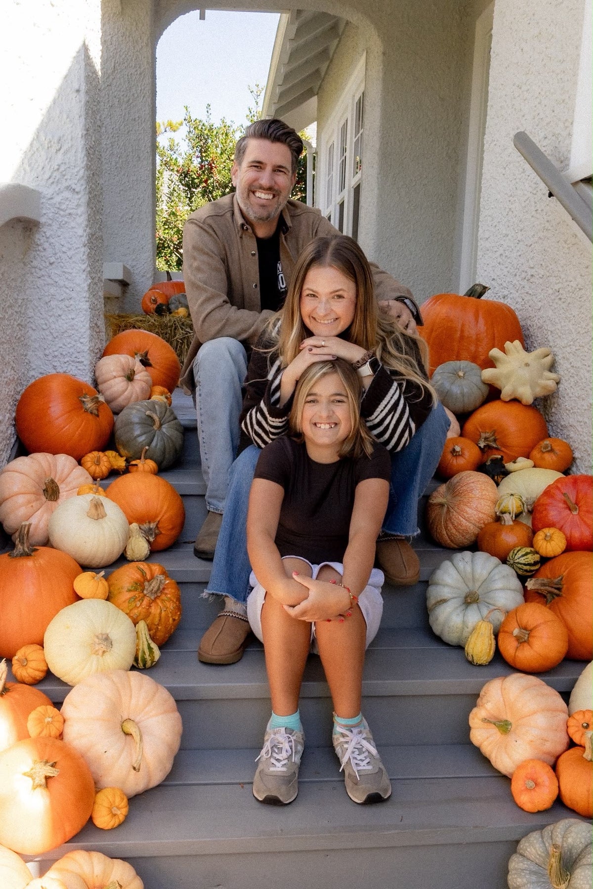 A family of three poses on the front steps of a house decorated with numerous pumpkins and gourds. The father sits behind his two daughters, who are smiling and posing together.