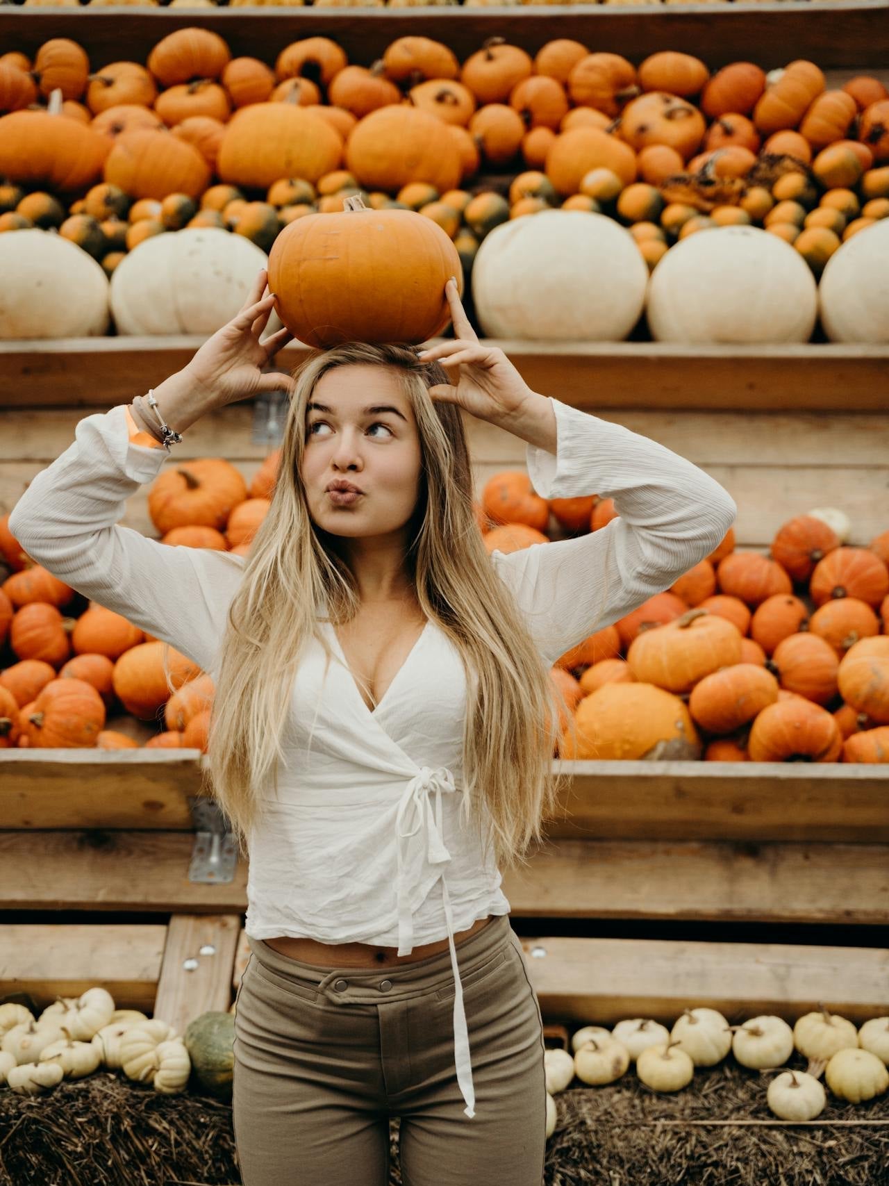 A woman with long blonde hair in a white wraparound shirt balances a pumpkin on her head in front of a display of pumpkins.