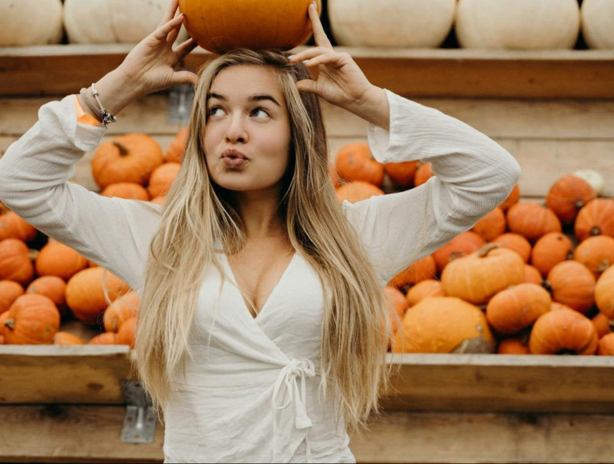 A woman with long blonde hair in a white wraparound shirt balances a pumpkin on her head in front of a display of pumpkins.