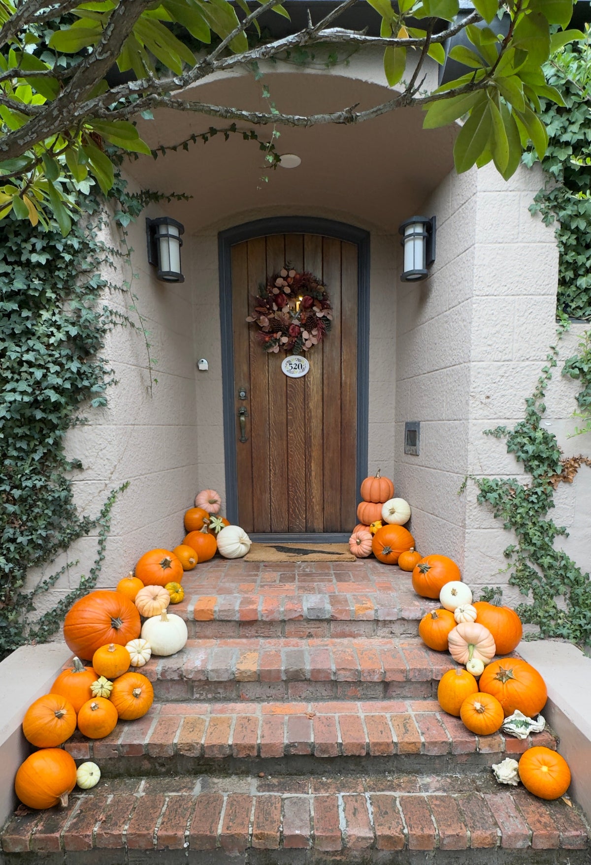A front porch with a brick staircase decorated with numerous orange, yellow, and white pumpkins and gourds leading up to a wooden door with a fall wreath.