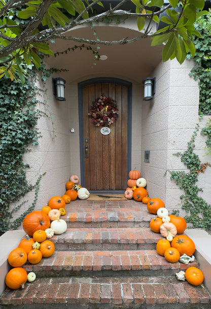 A front porch with a brick staircase decorated with numerous orange, yellow, and white pumpkins and gourds leading up to a wooden door with a fall wreath.