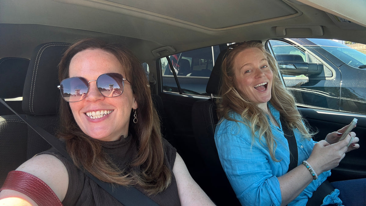 Two women smiling inside a car, with the woman on the left wearing sunglasses and the woman on the right wearing a bright blue shirt and holding a phone.