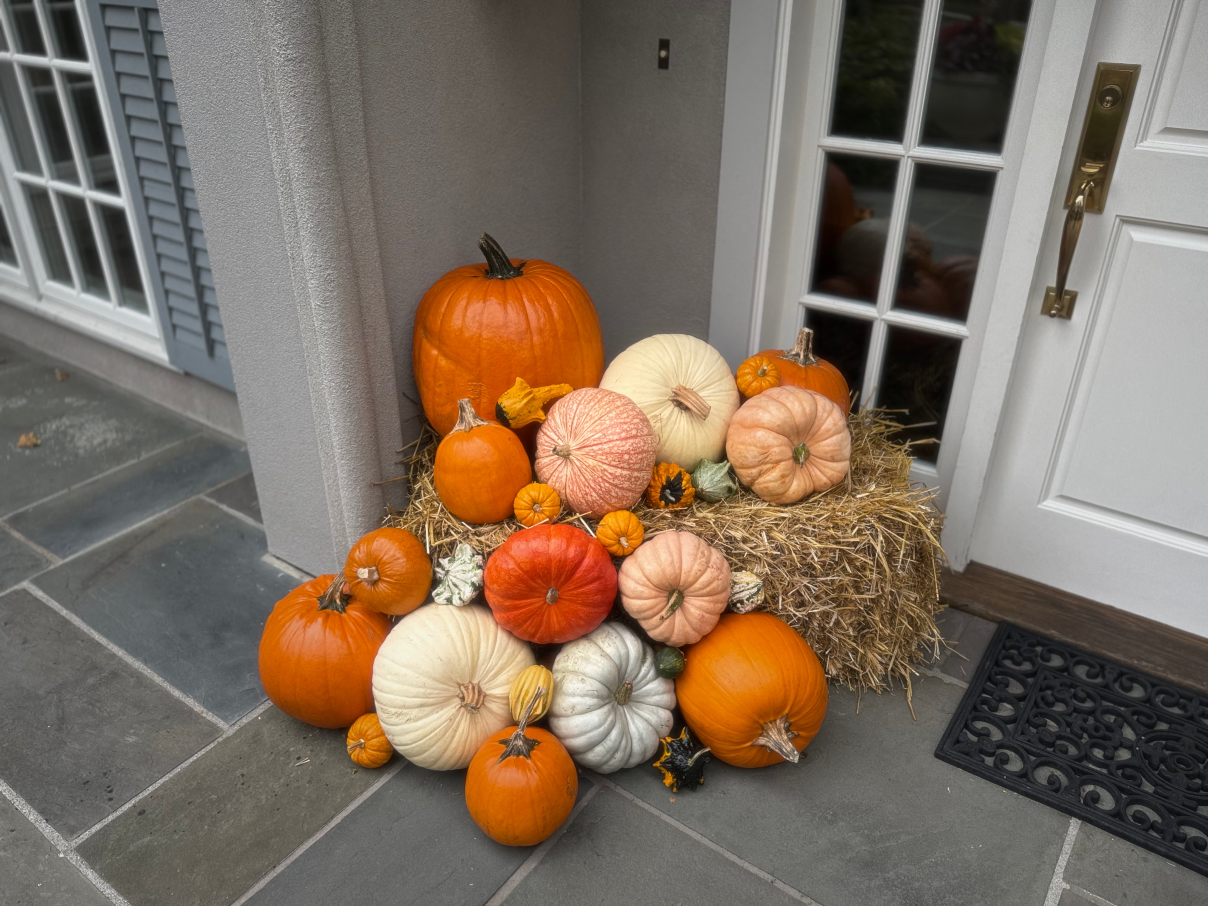 A decorative autumn pumpkin arrangement on a front porch, featuring multiple pumpkins and gourds in orange, cream, peach, and white placed on a bale of hay beside a white door.