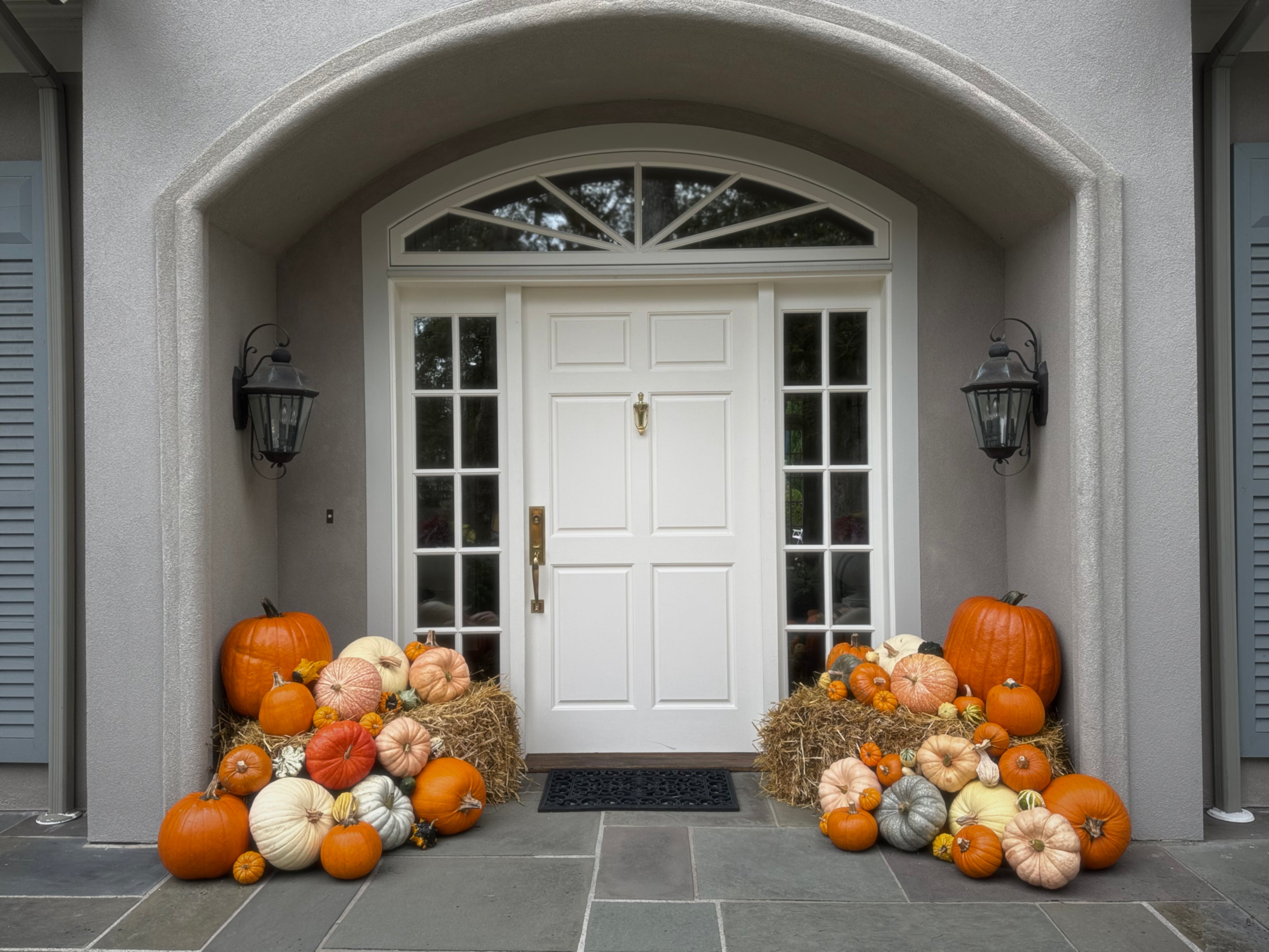 A front porch entryway with a white double door under a stone archway, decorated with numerous pumpkins and gourds arranged on hay bales and the stone steps.