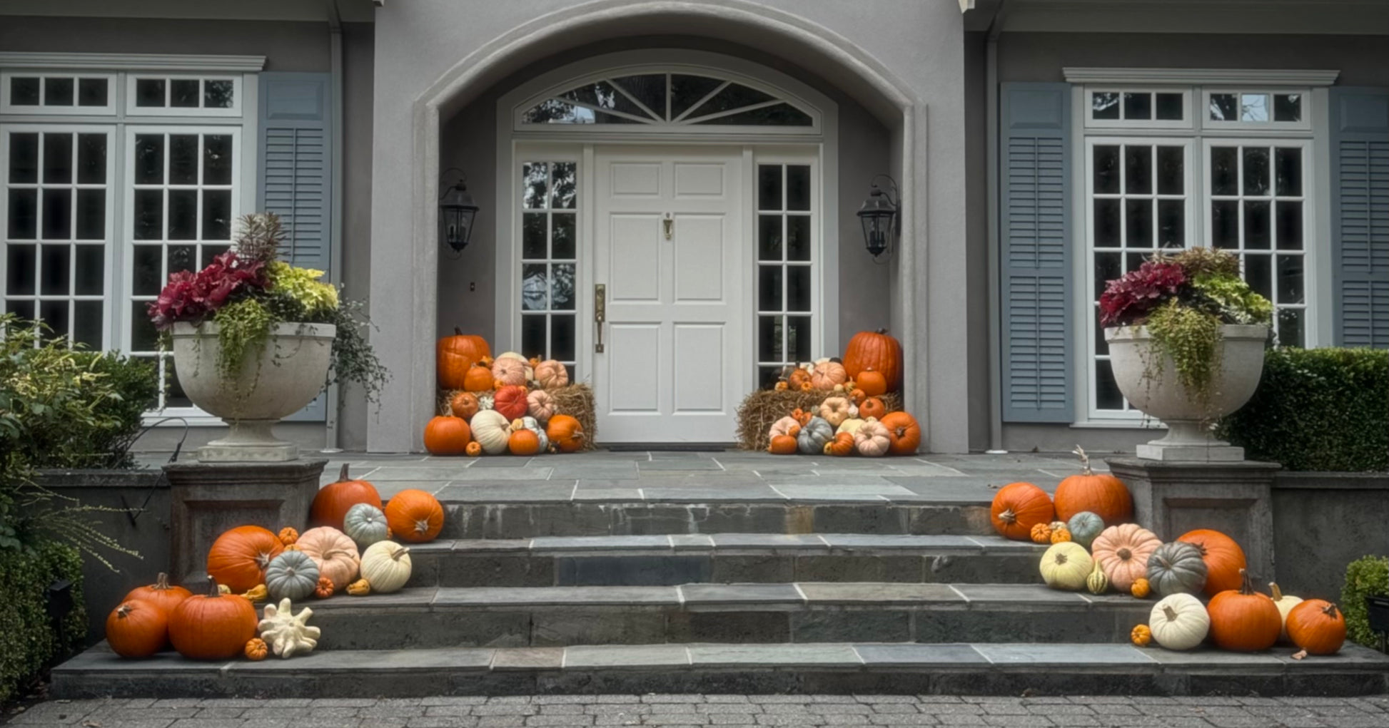 A front porch decorated with numerous pumpkins and gourds arranged on the steps and beside the white double doors, with large planters holding fall flowers on both sides.