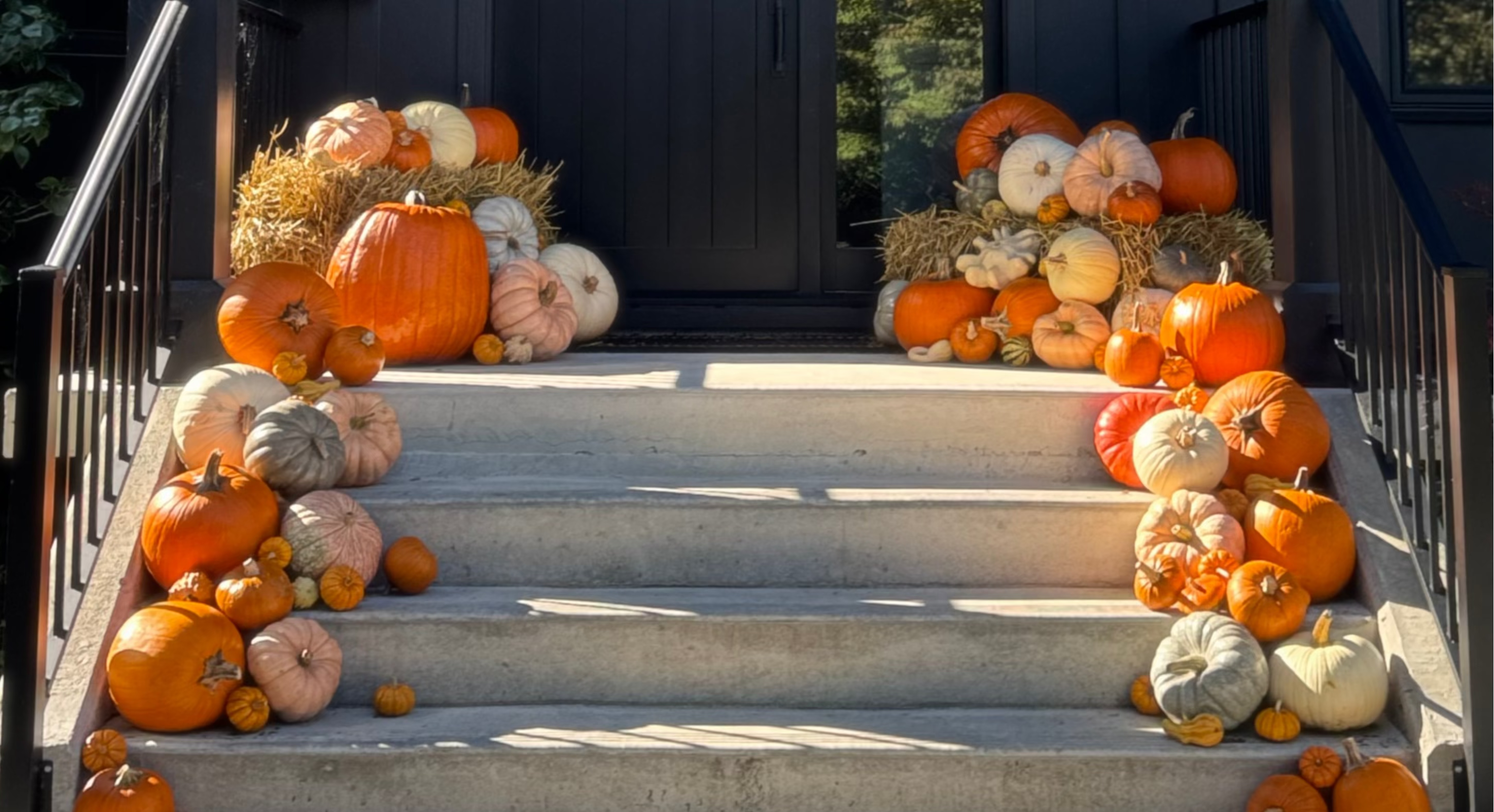 A decorative arrangement of assorted pumpkins and gourds in orange, beige, white, and muted pink tones, styled on a set of concrete steps leading up to a dark front door. Hay bales and black railings frame the scene.
