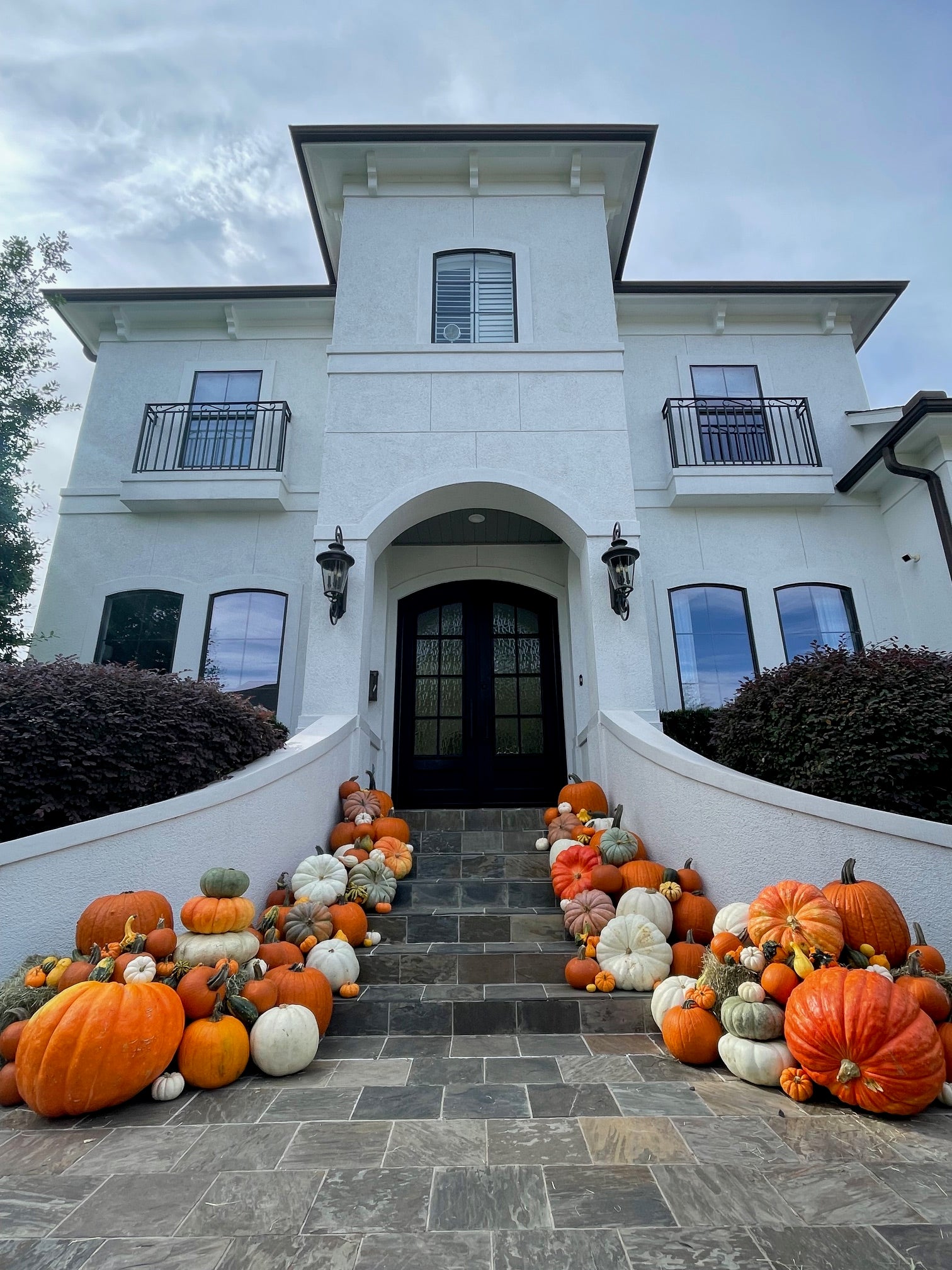A huge display of pumpkins of various shapes, sizes and colours sit atop three hay bales on a porch of a very large white house. 