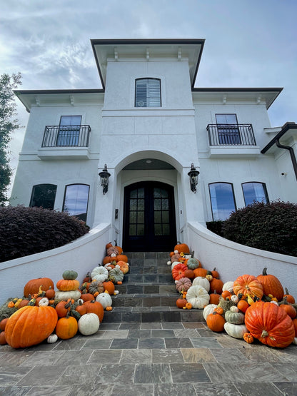 A huge display of pumpkins of various shapes, sizes and colours sit atop three hay bales on a porch of a very large white house. 