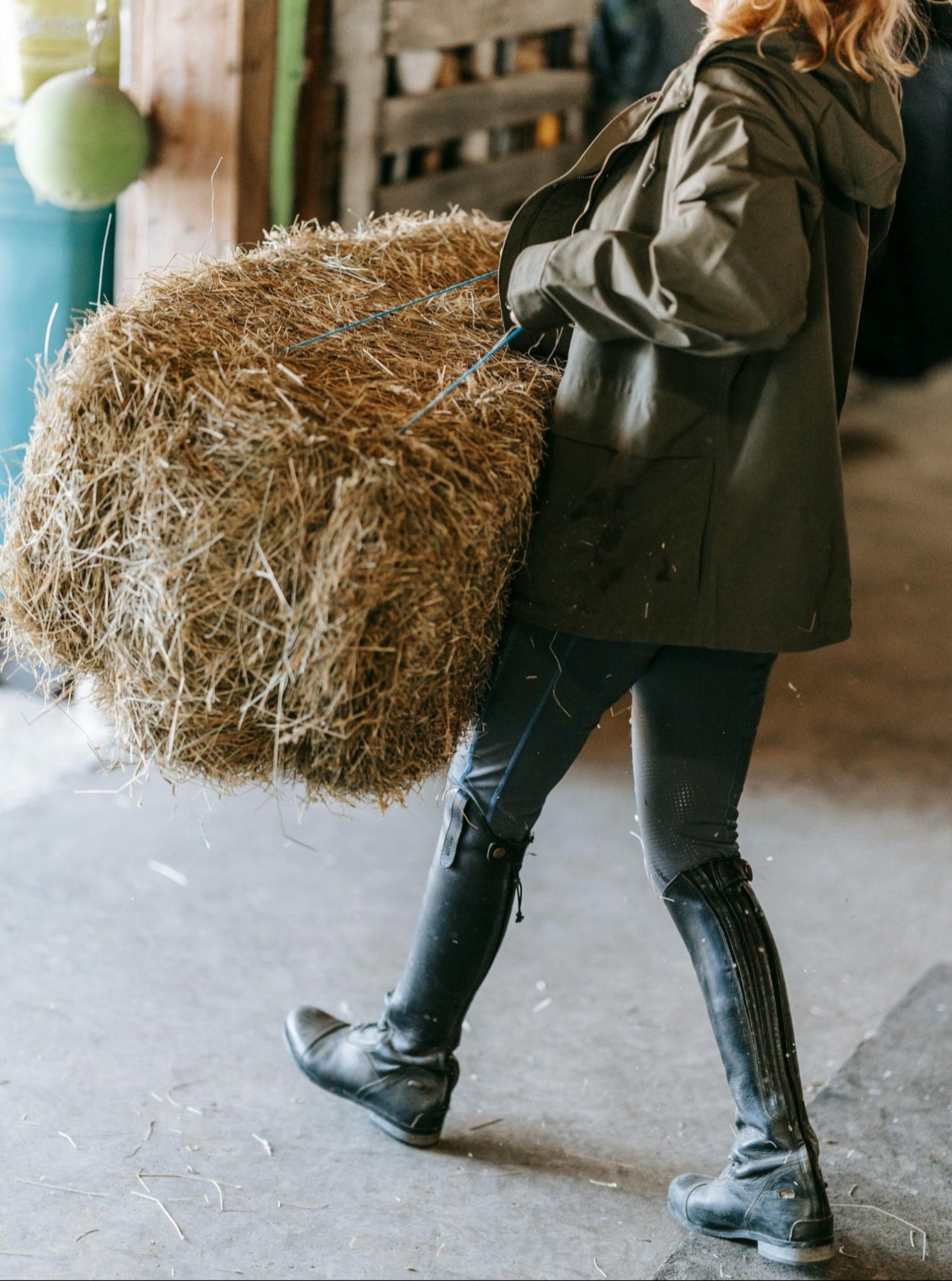A woman in tall boots and a raincoat carries a hay bale out of a barn.